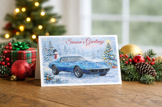 Season’s Greetings Christmas card featuring a 1968 blue Corvette in a snowy winter scene, displayed on a wooden table surrounded by festive decorations including wrapped gifts, ornaments, pinecones, greenery, and soft Christmas lights.