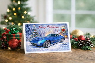 Christmas greeting card featuring a 1969 Corvette Stingray in a snowy winter scene, displayed on a wooden table surrounded by festive decorations including ornaments, pinecones, greenery, and soft Christmas lights