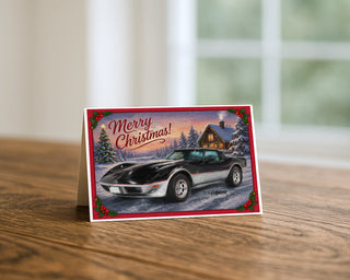 1978 Corvette Christmas greeting card standing on a wooden table beside a softly blurred window, showing the classic black and silver C3 in a snowy cabin scene with holly borders and Merry Christmas lettering. 5x7 holiday card by Will Davis Studios.