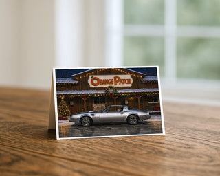 1979 Pontiac Trans Am Christmas greeting card standing on a wooden table beside a softly blurred window, showing the silver muscle car outside a snow-dusted country store with holiday lights and garland. 5x7 holiday card by Will Davis Studios.