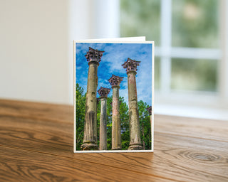 Will Davis Studios 5x7 fine art architecture greeting card featuring historic stone columns and ruins against a blue sky, displayed standing on a wooden table with a white envelope.