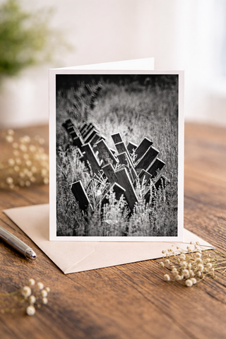 Black-and-white fine art photography greeting card featuring abstract wooden fence posts and tall grasses, displayed as a 5x7 folded card with envelope on a rustic tabletop.
