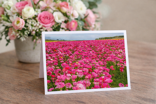 Floral card with a field of pink flowers on a wooden surface, next to a bouquet of pink and white flowers.