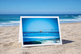 Print of a beach scene with a person in the water, placed on sand with ocean and sky in the background.