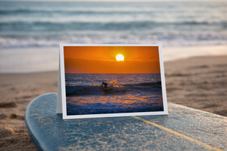 Sunset over water with a person on a surf board displayed on a beach.