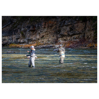 Two people fly fishing in a river with rocky cliffs in the background