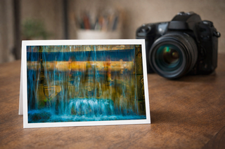 Greeting card with a watercolor design of a waterfall on a wooden surface next to a camera.