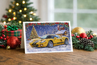 2005 Ford GT Christmas greeting card displayed on a wooden table with festive decorations including red ornaments, plaid gift box, pinecones, evergreen garland, and a softly lit Christmas tree in the background