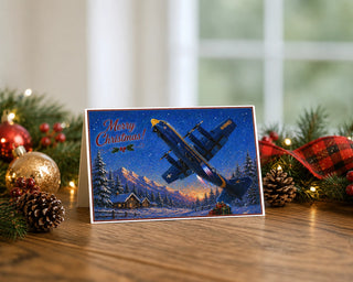 Blue Angels Fat Albert C-130 Christmas card displayed on a rustic wooden table with holiday decorations including pine garland, red and gold ornaments, pinecones, buffalo plaid ribbon, and warm twinkle lights. The 5x7 card features the Marines' navy and gold Hercules climbing over a snowy mountain cabin scene with Merry Christmas lettering.