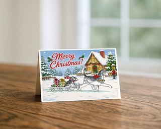 Beagle Christmas greeting card standing on a wooden table beside a softly blurred window, showing a cartoon beagle pulling a sled of presents and a puppy past a snowy cottage. 5x7 holiday card by Will Davis Studios.