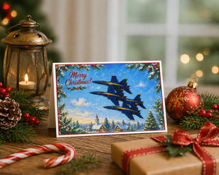 Blue Angels Christmas 5x7 greeting card from Will Davis Studios displayed on a wooden table with a brass lantern, pinecones, red ornament, candy cane, wrapped gift, and evergreen sprigs — three US Navy F/A-18 Hornets over a snowy village with Merry Christmas script, naval aviation holiday fine art card