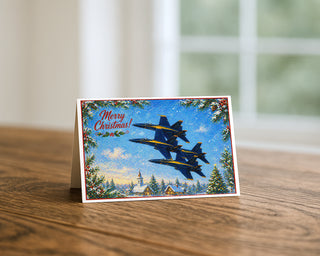 Blue Angels Christmas 5x7 greeting card from Will Davis Studios displayed standing on a wooden table near a bright window — four US Navy F/A-18 Hornets in stepped formation over a snowy village with church steeple, holly border and Merry Christmas script, naval aviation holiday fine art card