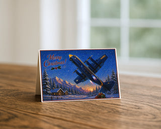 Blue Angels Fat Albert Christmas card displayed on a wooden table, featuring the Marine Corps C-130 flying over a snowy mountain cabin scene with “Merry Christmas” text.