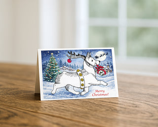 7x5 landscape Christmas greeting card displayed on a wooden table featuring a whimsical illustration of a white dog Welsh Corgi wearing reindeer antlers and carrying a gift in a snowy holiday village scene with a Christmas tree and the message “Merry Christmas!”.