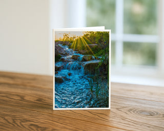 Desert stream at sunrise fine art landscape photography greeting card standing on a wooden table, featuring golden sun rays over flowing water and rocks in a Southwest nature scene by Matt Punches from Will Davis Studios.