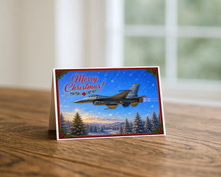 F-16 Fighting Falcon Christmas greeting card standing on a wooden table beside a softly blurred window, showing the fighter jet over a snowy landscape with a lit Christmas tree, holly corners, and Merry Christmas lettering. 5x7 holiday card by Will Davis Studios.