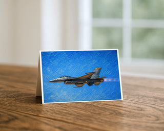 F-16 Fighting Falcon greeting card standing on a wooden table beside a softly blurred window, showing the painterly fighter jet in flight with afterburner glowing against a textured brushstroke blue sky. 5x7 aviation art card by Will Davis Studios.