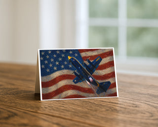 Fat Albert C-130 American flag 5x7 greeting card from Will Davis Studios displayed standing on a wooden table near a bright window — US Marine Corps C-130 Hercules in Blue Angels colors during powered climb against painterly American flag, Marine aviation patriotic fine art card