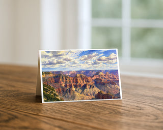 Grand Canyon watercolor 5x7 greeting card from Will Davis Studios displayed standing on a wooden table near a bright window — layers of red and gold sandstone buttes fading to lavender at the horizon under a wide Arizona sky, Southwest landscape fine art card