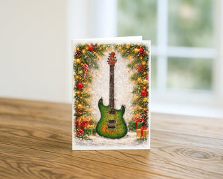 Green electric guitar Christmas greeting card displayed on a wooden table with festive garland, ornaments, lights, and wrapped holiday gifts, music-themed holiday card by Will Davis Studios.