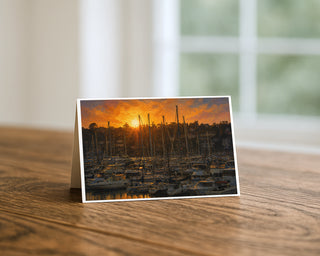 Harbor sunset marina greeting card standing on a wooden table beside a softly blurred window, showing painterly sailboats at golden hour with masts silhouetted against a glowing orange sky. 5x7 fine art card by Will Davis Studios.