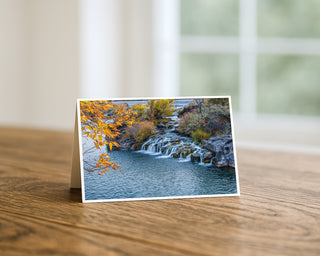 7x5 Idaho Falls autumn waterfall fine art photography greeting card displayed on a wooden table, featuring colorful fall foliage and cascading water by Will Davis Studios