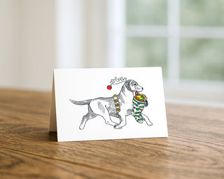 Illustration of a Labrador Retriever wearing reindeer antlers and a festive harness, carrying a Christmas stocking filled with toys on a holiday greeting card displayed on a wooden table. Will Davis Studios