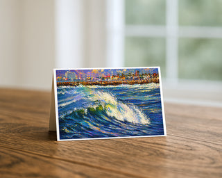 Ocean Beach California 5x7 greeting card from Will Davis Studios displayed standing on a wooden table near a bright window — painterly cresting wave with palm-lined San Diego shoreline and jetty, coastal fine art card