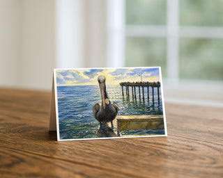 Ocean Beach pier and pelican watercolor 5x7 greeting card from Will Davis Studios displayed standing on a wooden table near a bright window — brown pelican perched on a piling with the Ocean Beach pier and fishermen in the background, coastal fine art card