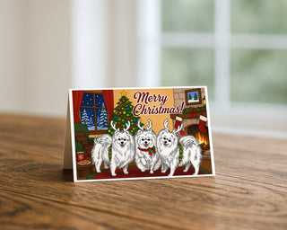 Pomeranian Christmas greeting card standing on a wooden table beside a softly blurred window, showing three cartoon Pomeranians in reindeer antlers beside a Christmas tree and fireplace. 5x7 holiday card by Will Davis Studios.