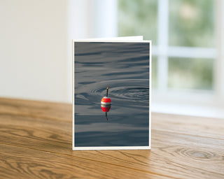 Red fishing bobber floating on calm water with circular ripples on a lake, minimalist nature photography greeting card by Will Davis Studios.