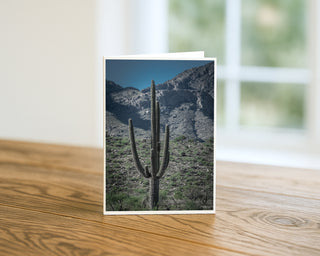 Tall saguaro cactus standing in the Arizona desert with mountain backdrop, featured on a vertical photography greeting card displayed on a wooden table.