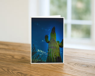 5x7 greeting card featuring a dramatic Saguaro cactus against a deep blue Arizona sky, displayed upright on a wooden table with soft natural light background