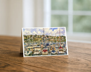San Diego harbor marina watercolor 5x7 greeting card from Will Davis Studios displayed standing on a wooden table near a bright window — sailboats and cabin cruisers in their slips with Point Loma hillside and red-tile rooftops behind, coastal fine art card