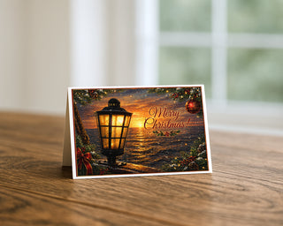 Star of India lamp Christmas 5x7 greeting card from Will Davis Studios displayed standing on a wooden table near a bright window — glowing brass-and-glass ship lantern at sunset with snowfall, holly border, red ornament, velvet bow, and Merry Christmas script, maritime holiday fine art card