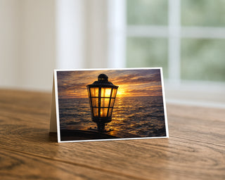 Star of India lamp at sunset 5x7 greeting card from Will Davis Studios displayed standing on a wooden table near a bright window — glowing brass-and-glass ship lantern with golden Pacific sunset and water reflection, maritime fine art card