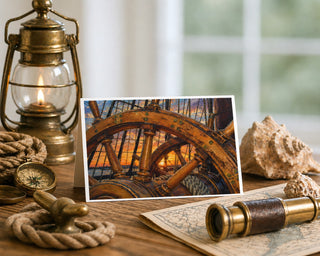 Star of India ship's wheel at sunset 5x7 greeting card from Will Davis Studios displayed on a wooden table with a brass lantern, vintage compass, telescope, antique map, seashells, and coiled rope — historic tall ship helm at golden Pacific sunset, maritime fine art card