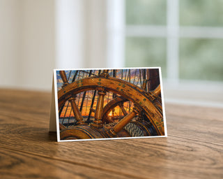 Star of India ship's wheel at sunset 5x7 greeting card from Will Davis Studios displayed standing on a wooden table near a bright window — historic tall ship's wooden helm with brass-banded spokes against golden Pacific sunset and rigging, maritime fine art card