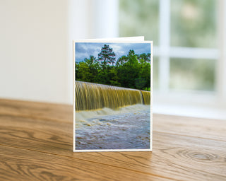 Starr's Mill Falls Georgia 5x7 greeting card from Will Davis Studios displayed standing on a wooden table near a bright window — Southern waterfall cascading over historic dam with green trees and stormy sky, landscape fine art card