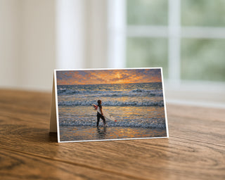 Surfer at sunset greeting card standing on a wooden table beside a softly blurred window, showing a painterly scene of a lone surfer in the shallows with golden waves and sunset reflections. 5x7 fine art card by Will Davis Studios.