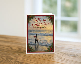 Surfer Christmas greeting card standing on a wooden table beside a softly blurred window, showing a surfer in the shallows at golden hour framed by crimson borders, holly, and Merry Christmas lettering. 5x7 holiday card by Will Davis Studios.