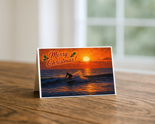 Surfing sunset Christmas greeting card standing on a wooden table beside a softly blurred window, showing a surfer riding a wave under a blazing orange sky with Merry Christmas lettering and holly. 5x7 holiday card by Will Davis Studios.