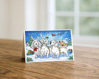Illustrated Christmas card featuring three dogs wearing reindeer antlers in a snowy winter scene with decorated tree, gifts, lights, and Santa’s sleigh in the sky, displayed on a wooden table.