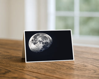 Waning gibbous moon 5x7 greeting card from Will Davis Studios displayed standing on a wooden table near a bright window — detailed lunar surface with craters and shadowed terminator against a deep blue-black night sky, astrophotography fine art card