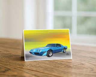 Will Davis Studios 5x7 greeting card featuring an illustration of a blue 1979 Pontiac Trans Am classic muscle car on a yellow sunset gradient background, displayed standing on a wooden table