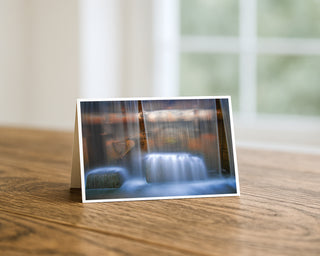Product mockup of a Will Davis Studios 5x7 fine art greeting card in landscape orientation standing upright on a warm wooden table, featuring a breathtaking long exposure photograph of Arizona Falls shot from behind the waterfall with silken blue-white curtains of cascading water framing warm amber and rust-toned historic cast iron hardware and weathered stonework beyond, photographed in soft natural light with a bright window in the background.