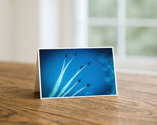 Product mockup of a Will Davis Studios 5x7 fine art greeting card in landscape orientation standing upright on a warm wooden table, featuring six Blue Angels jets performing a dramatic fan break with white smoke trails against a deep cobalt blue sky, with soft natural window light in the background.