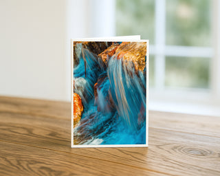 Will Davis Studios 5x7 fine art nature greeting card featuring a vibrant blue waterfall cascading over textured rocks, displayed standing on a wooden table