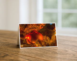 Product mockup of a Will Davis Studios 5x7 fine art greeting card in landscape orientation standing upright on a warm wooden table, featuring a glowing red Christmas ornament nestled in golden-lit evergreen branches with warm amber holiday lights, photographed in soft natural light with a bright window in the background.