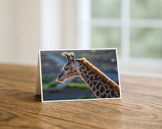 Product mockup of a Will Davis Studios 5x7 fine art greeting card in landscape orientation standing upright on a warm wooden table, featuring a stunning close-up profile portrait of a young giraffe with richly patterned neck, glowing amber mane, and wild dark hair tuft against a soft blue and green bokeh background, photographed in soft natural light with a bright window in the background.